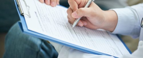 Crop shot of anonymous doctor in white gown writing with pen on clipboard during home visit